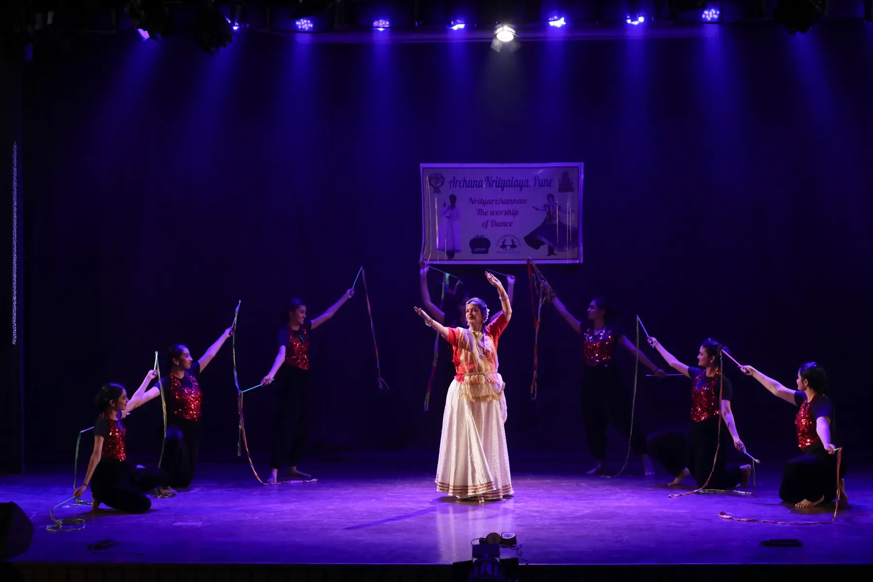 Guru Archana Patwardhan centre stage under violet light, flanked by dancers, at the Nrityarchannaa annual gathering in Pune