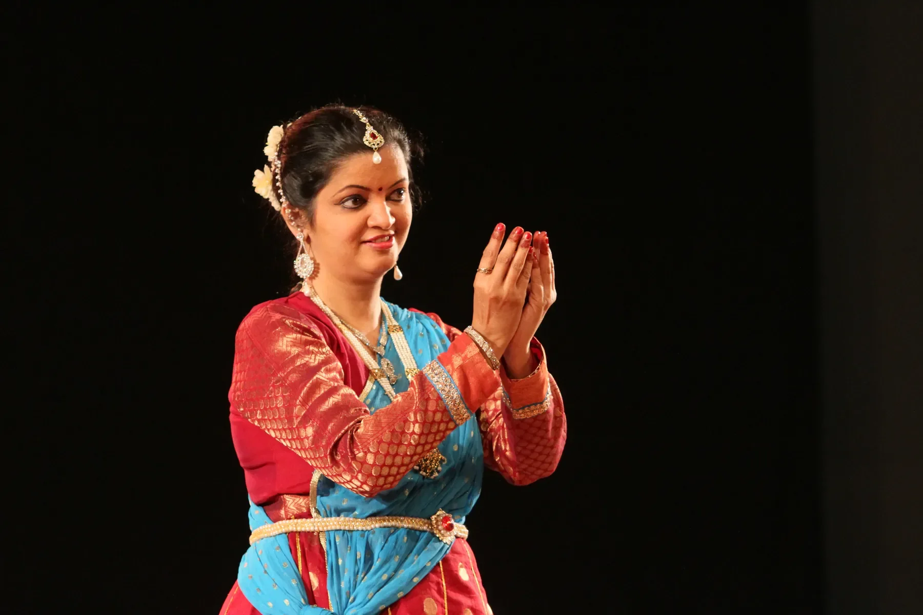 Guru Archana Patwardhan holding a namaskar hastak in red and turquoise on a darkened stage