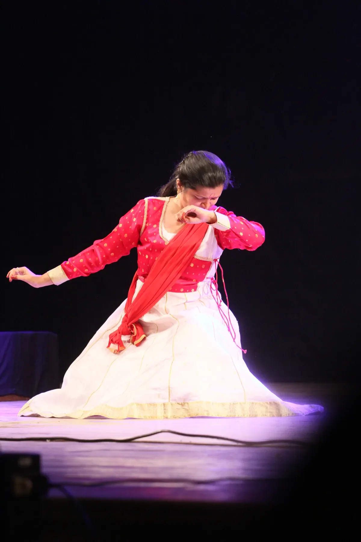 Guru Archana Patwardhan kneeling in a pool of light on stage, drawing a red dupatta across her arm
