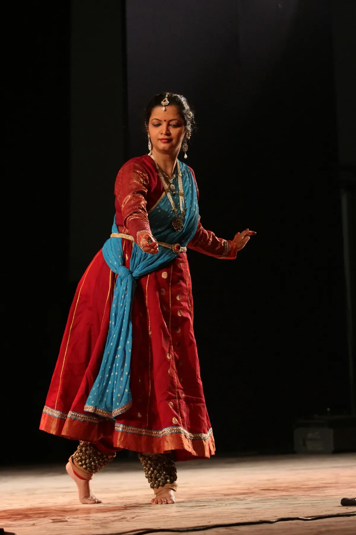 Guru Archana Patwardhan mid-phrase in a maroon and turquoise Kathak costume, ghungroos at the ankle