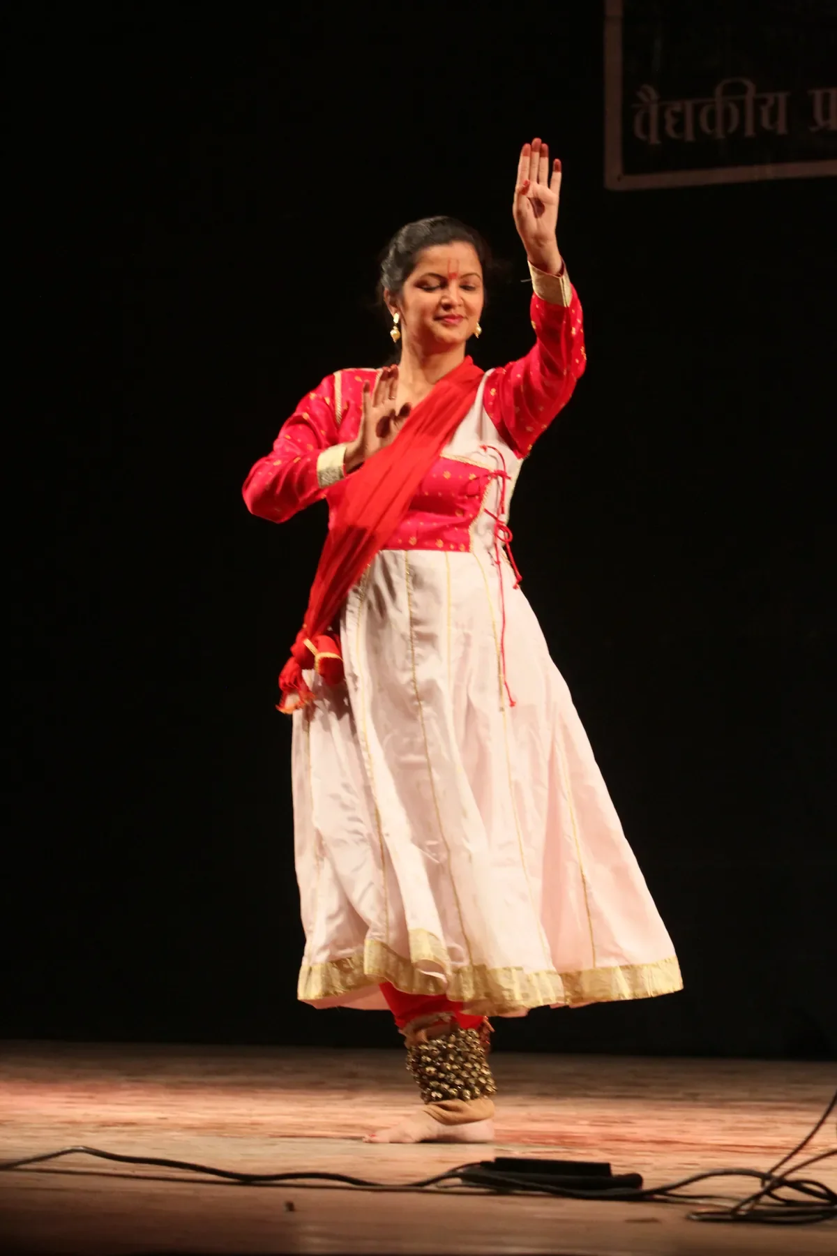 Guru Archana Patwardhan standing in a red and white Kathak costume, right hand raised in a hastak